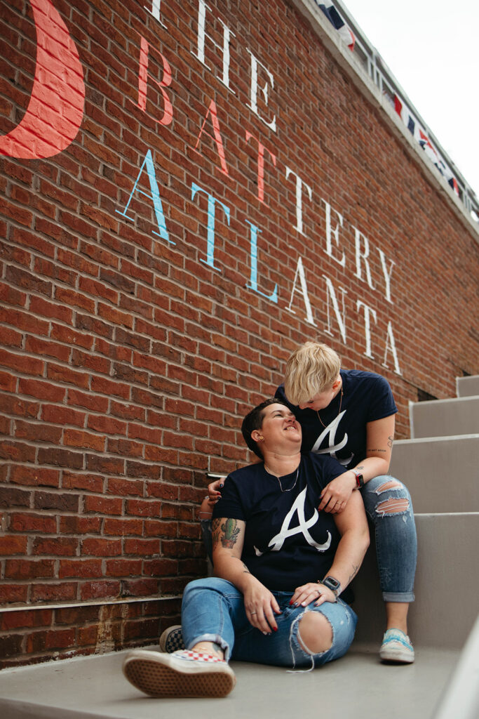This photo shows a queer couple after their Atlanta engagement cuddling while sitting under a sign that says The Battery Atlanta
