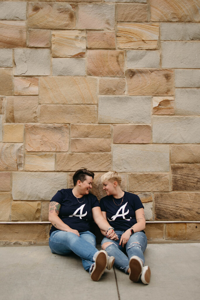 This photo shows a queer couple sitting side by side on the ground holding hands after their Atlanta engagement. They are both wearing Atlanta Braves shirts.