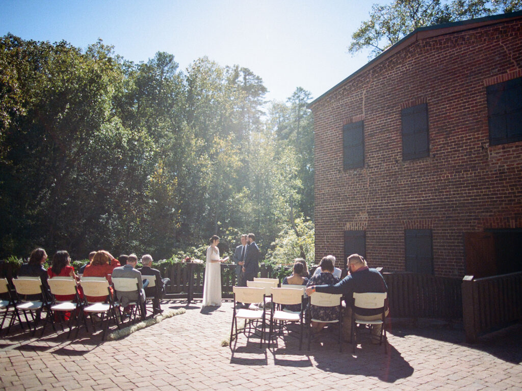 Roswell Mill wedding day ceremony captured on film. Shows couple at the front of the ceremony with the sun shining down.